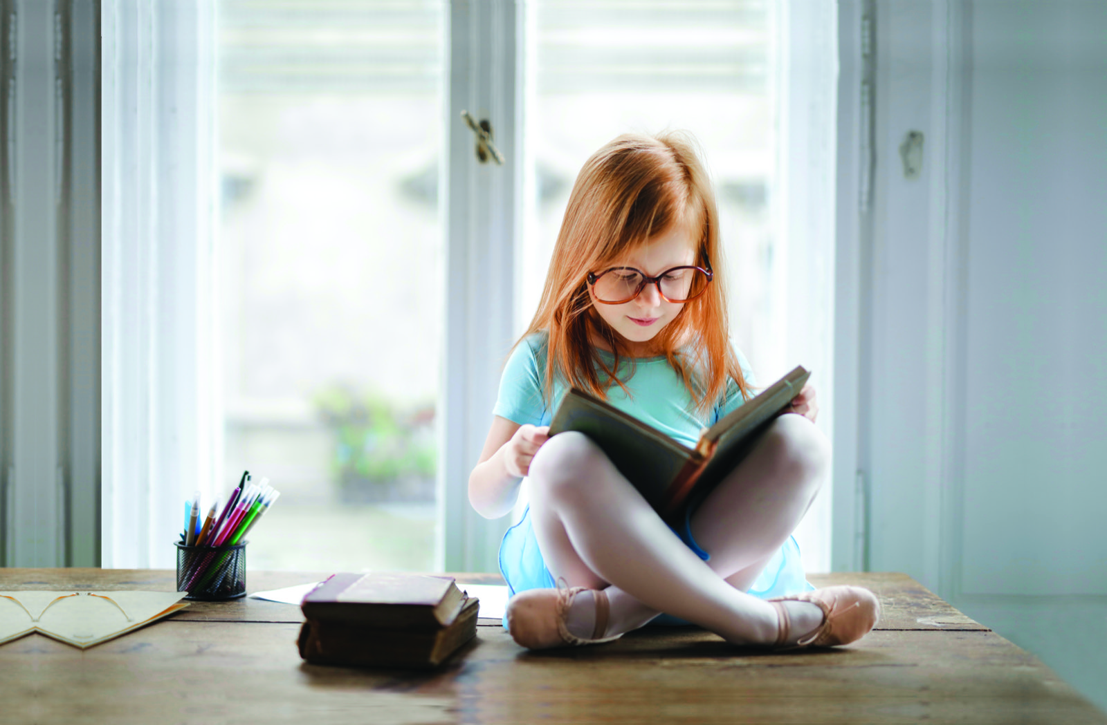 A young girl in glasses sits cross-legged on a wooden table by a bright window, absorbed in reading a book.